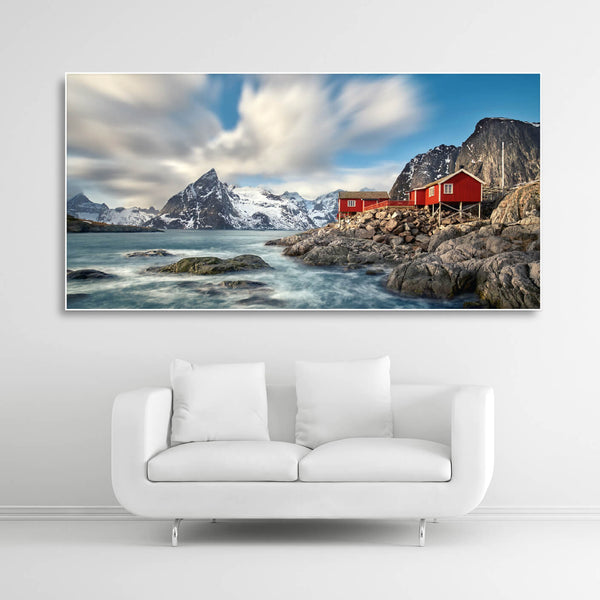 Schallschutzbild mit Felsen, Wasser und roter Holzhütte. Weißer Rahmen im Querformat über weißem Sofa.