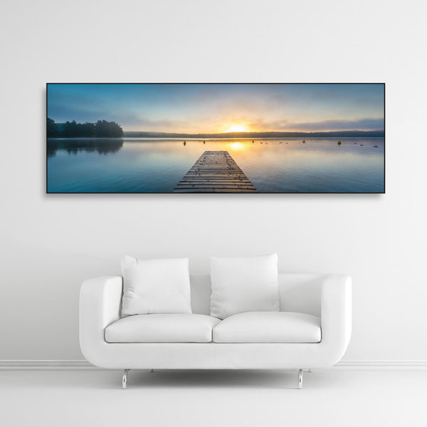 Schallschutzbild mit Bootssteg im See bei Sonnenaufgang. Schwarzer Rahmen im Querformat über weißem Sofa.