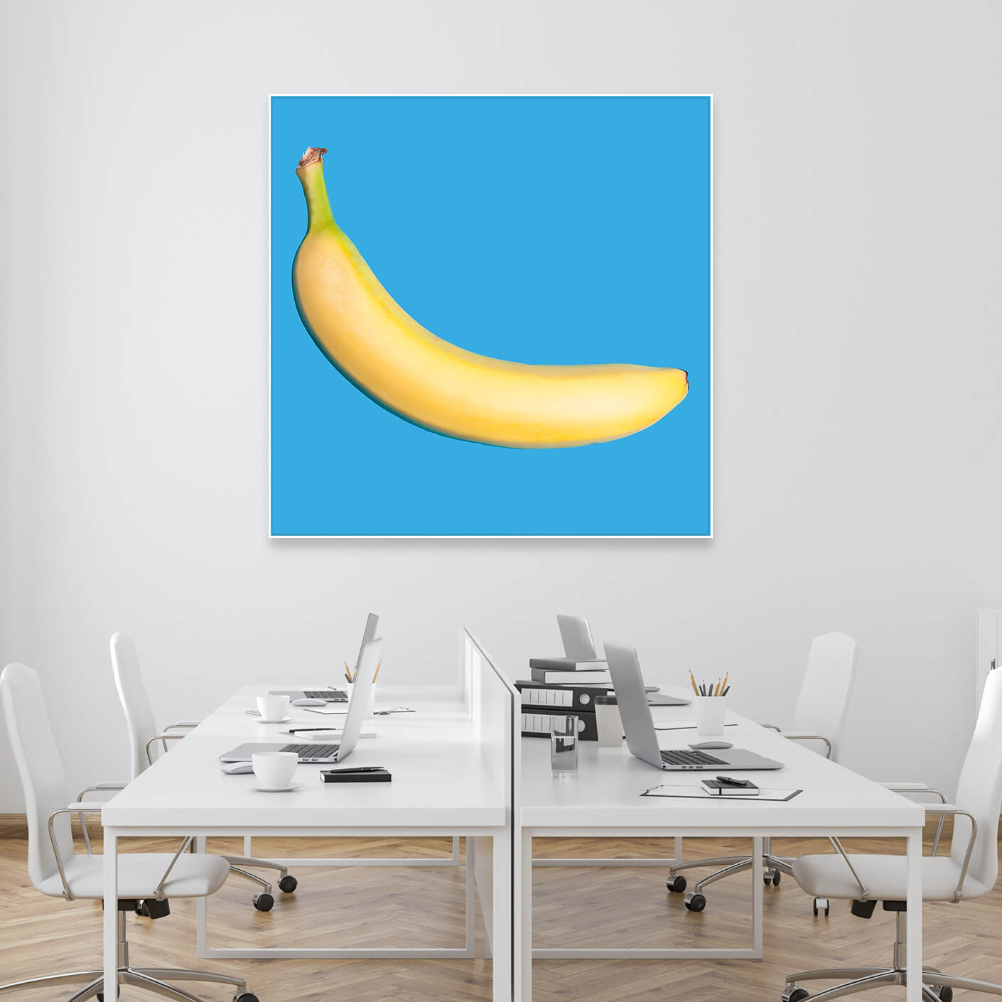 Modernes Loft mit mehreren Schreibtischen und Tysta Akustikbild mit Fotografie einer gelben Banane auf hellblauen Hintergrund. Weißer Rahmen im Quadrat.