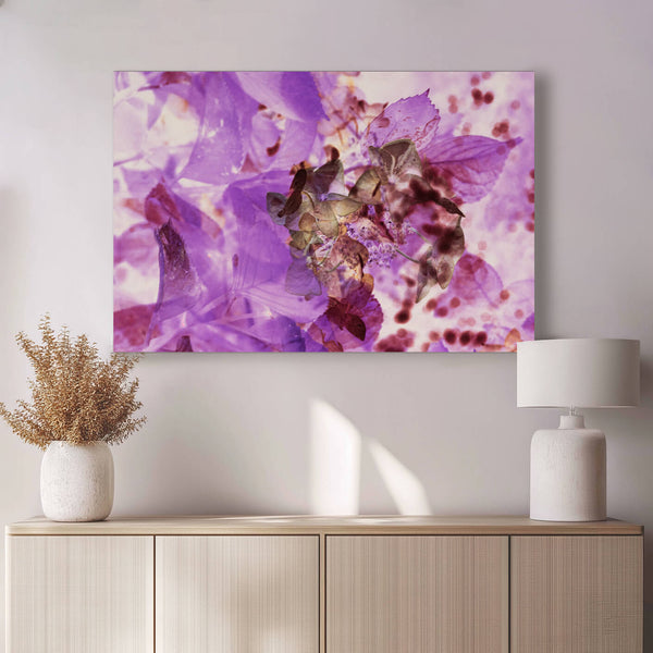 Ein abstraktes Gemälde in verschiedenen Violetttönen mit floralen Anklängen hängt über einer hellen Kommode.  Warmes Sonnenlicht fällt von rechts auf das Bild und die Kommode, auf der eine weiße Vase mit getrockneten Gräsern und eine Tischlampe stehen.