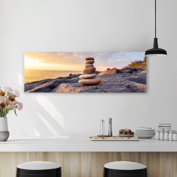 Ein Panorama-Wandbild mit einem Stapel aus gestapelten Steinen am Strand bei Sonnenuntergang. Das Bild hängt über einer modernen Küchenzeile mit weißen Arbeitsplatten und Dekorationen wie Blumen und Gewürzen. Barhocker stehen an der Theke, was auf einen offenen Wohnbereich hindeutet. 