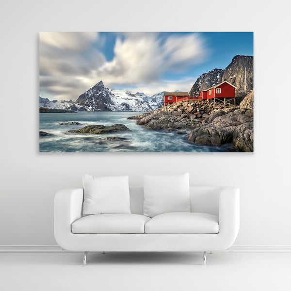 Ein großes Landschaftsbild zeigt eine raue Küstenlandschaft mit mehreren kleinen, roten Häusern, die auf Felsen am Meer gebaut sind. Im Hintergrund erheben sich schneebedeckte Berge unter einem dramatischen Himmel mit weißen Wolken. Bildformat 160x90cm.