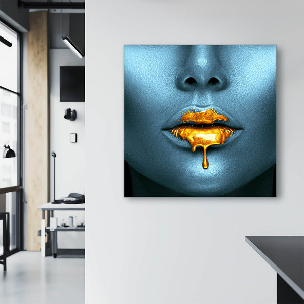 Ein großes Frauenportrait hängt an einer weißen Wand in einem modernen Atelier. Das Bild zeigt eine Nahaufnahme eines Frauengesichts in kühlen Blautönen. Die Lippen sind mit goldener Farbe bedeckt, die an den Mundwinkeln heruntertropft