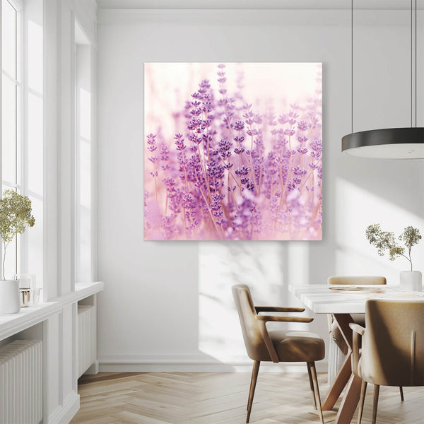 Eine große quadratische Fotografie hängt an einer weißen Wand in einem Esszimmer. Das Bild zeigt eine Nahaufnahme von Lavendelblüten in verschiedenen Violetttönen. Die Blüten sind leicht verschwommen und wirken verträumt. Im Hintergrund ist ein sanftes Licht zu sehen. Der Raum ist hell und modern eingerichtet mit einem Holztisch und beigen Stühlen.