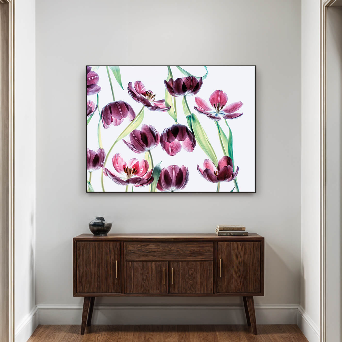 Ein elegantes Bild von mehreren Tulpen hängt an einer hellen Wand über einem braunen Sideboard. Die Tulpen haben zarte, violette Blütenblätter mit dunkleren Akzenten und grünen Stängeln. Sie scheinen vor einem hellen Hintergrund zu schweben, was ihnen eine ätherische Qualität verleiht. Das Sideboard trägt eine kleine Schale und ein paar Bücher.