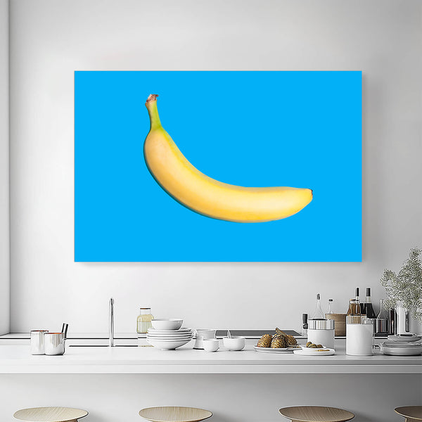 Pop-Art-Druck einer einzelnen Banane auf leuchtendem Blau. Das Bild hängt an einer weißen Wand über einer modernen Küchenzeile mit weißen Arbeitsplatten und Holzregalen. Die Küche ist hell und luftig mit minimalistischer Dekoration.