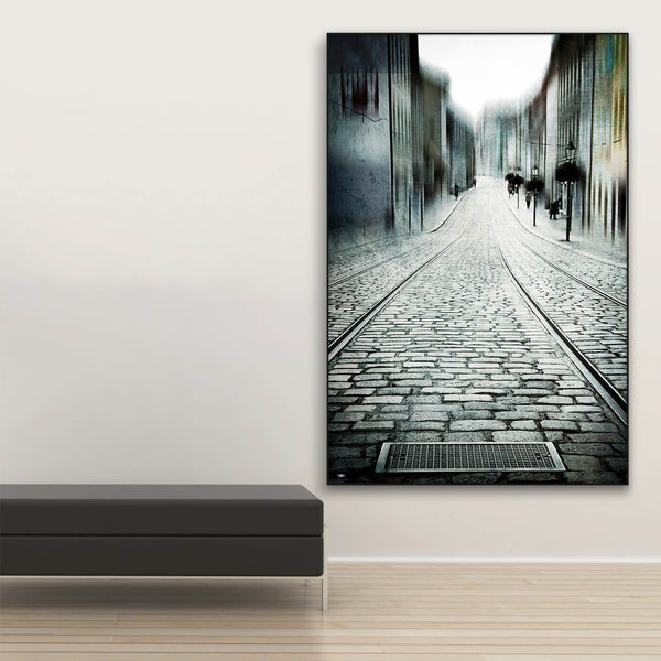 Tysta Akustikbild Old City von Christiane Steinicke. Gasse einer Altstadt mit historischem Kopfsteinpflaster und Straßenbahnschienen. Schwarzer Bilderrahmen im Hochformat neben Sitzbank.